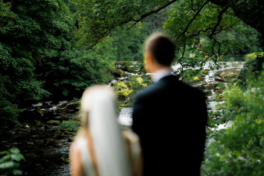 Soft focus analogue-style wedding photograph of a bride and groom standing beside a river beneath overhanging trees in the countryside.