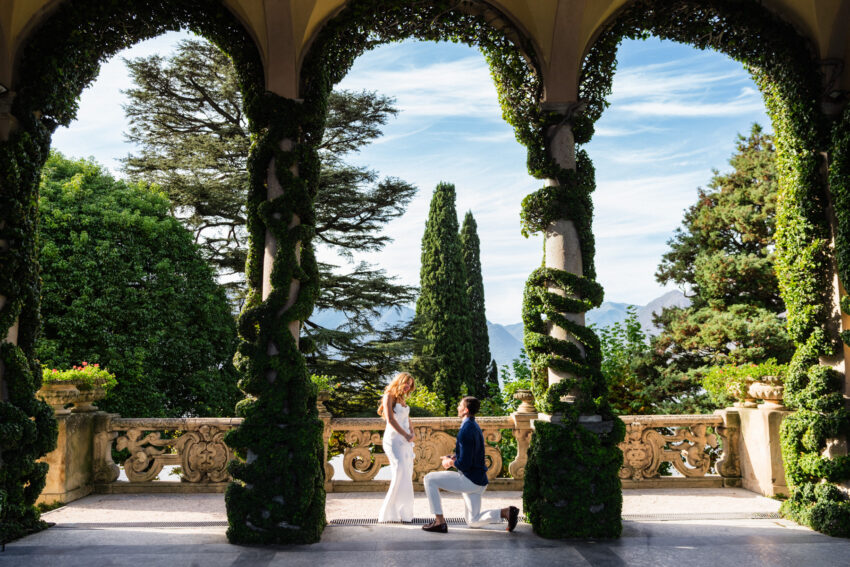 Elegant Lake Como proposal photograph beneath ivy-covered arches overlooking the Italian lakes and mountains.