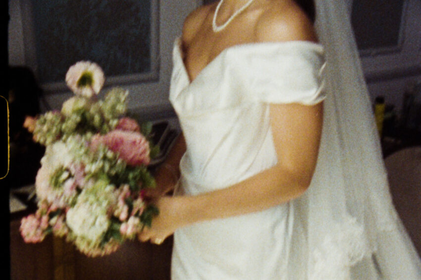 Grainy analogue wedding photograph of a bride holding a bouquet in a satin off-the-shoulder dress, captured with a soft vintage film aesthetic.