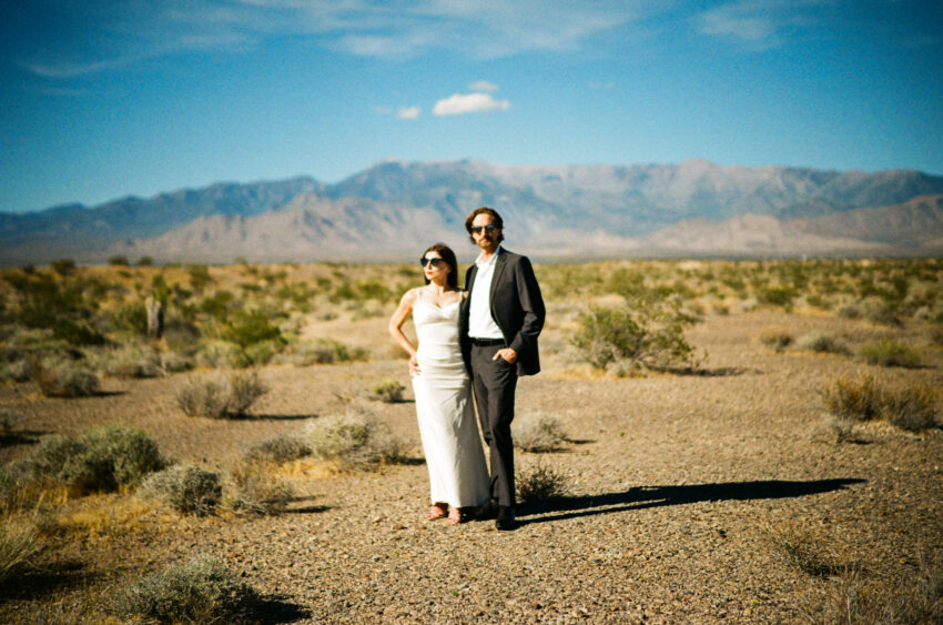 Stylish bride and groom standing in the Las Vegas desert with mountains in the background, photographed on film with soft focus and warm sunlight.