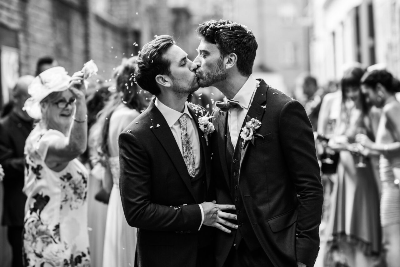 Black and white wedding photo of two grooms kissing after their ceremony as confetti falls around them in a city street, surrounded by guests. Elegant candid moment with soft background blur, emotional reportage-style same-sex wedding photography.
