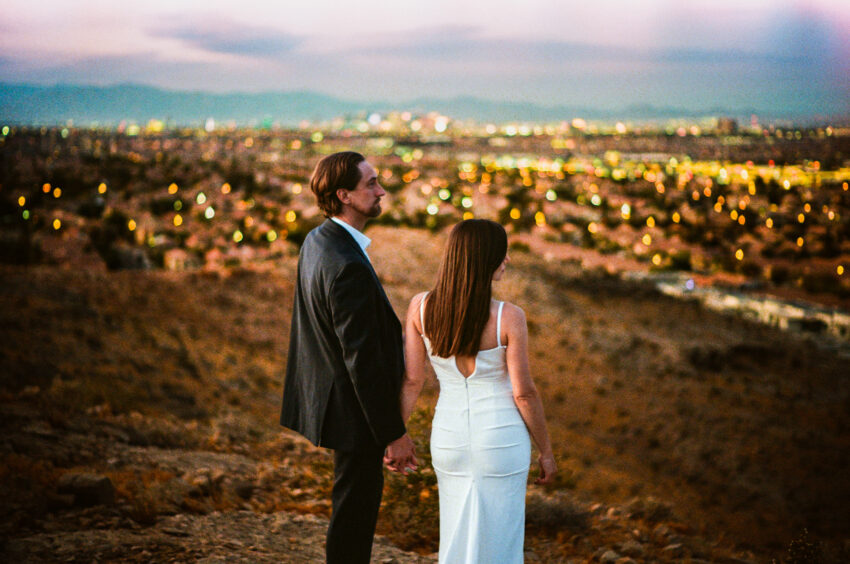 Bride and groom standing hand in hand on a hillside overlooking a city at dusk, city lights softly glowing in the distance, captured in colour on 35mm film with soft grain and a timeless feel.
