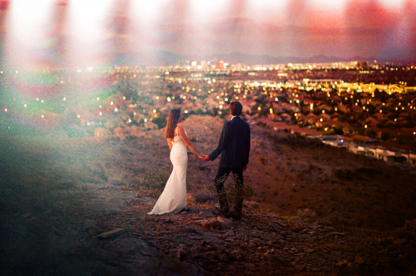 Bride and groom holding hands on a hillside overlooking Las Vegas at dusk, city lights glowing below, captured on 35mm film with warm tones, natural grain, and light leaks.