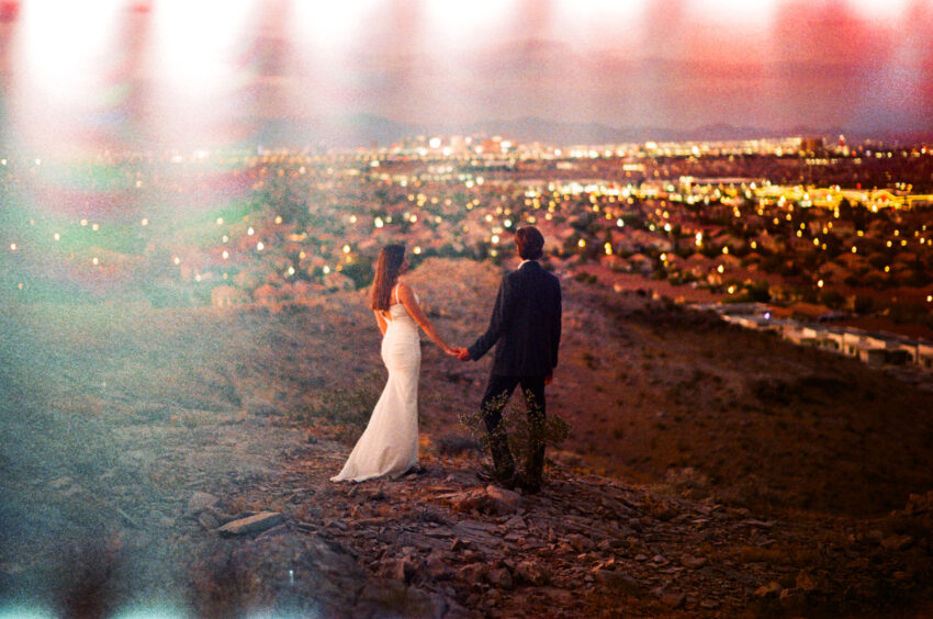 Bride and groom holding hands on a hillside overlooking Las Vegas at dusk, city lights glowing below, captured on 35mm film with warm tones, natural grain, and light leaks.