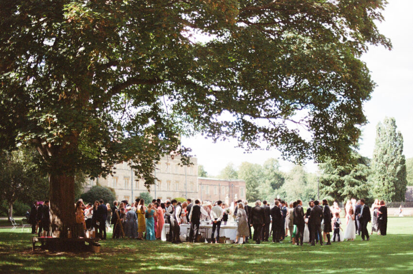 Guests gather beneath a large tree during an outdoor wedding reception, photographed on analogue 35mm film. The grainy image shows groups of people mingling on a lawn in front of a historic country house, with dappled sunlight filtering through the branches above.