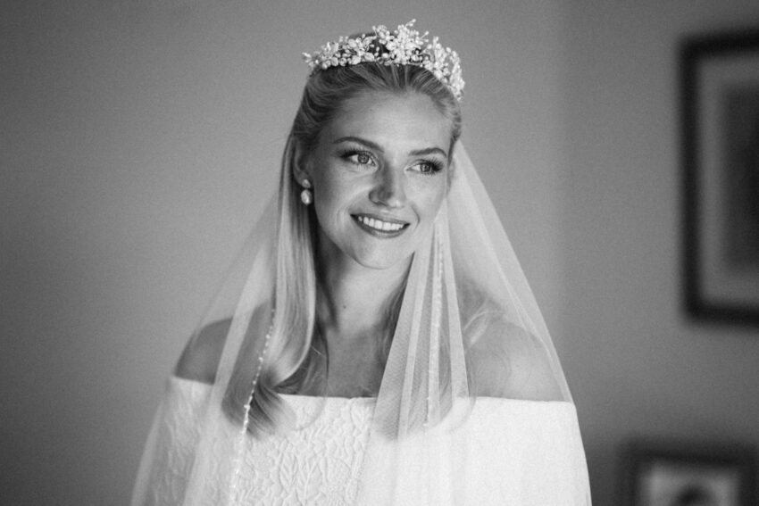 Bride wearing a floral tiara and veil, smiling softly beside a window. A black and white portrait with gentle light, fine grain, and an elegant, timeless feel.
