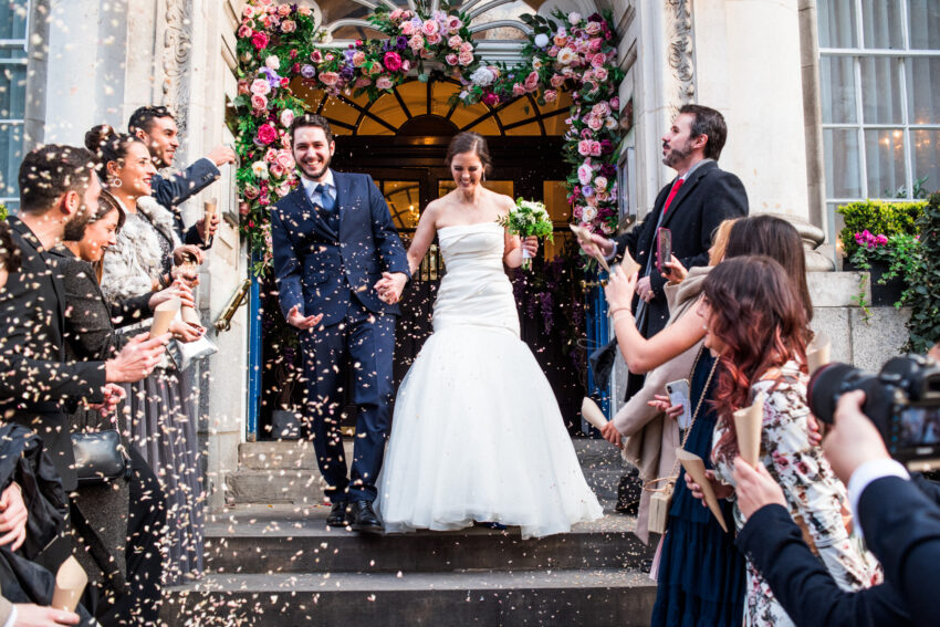 Bride and groom walking through a shower of confetti outside a flower-adorned wedding venue. Guests line the steps cheering as the couple smile and hold hands during their celebration.