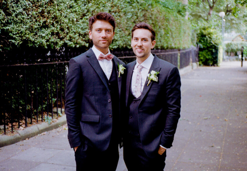 Two grooms standing together outdoors before their wedding, wearing dark suits with floral buttonholes. Film photograph with soft colours, shallow depth of field, and a relaxed portrait style.