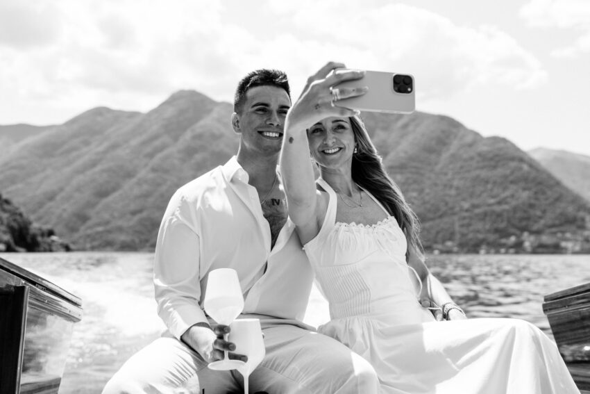 Couple taking a selfie on a wooden boat on Lake Como, smiling towards a smartphone with mountains and water in the background. Black and white image captured in bright sunlight.