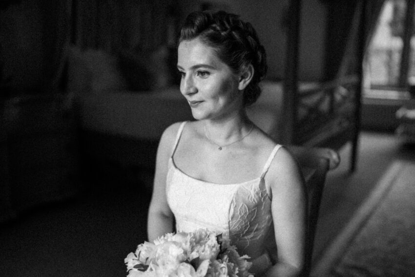 Bride seated indoors before the ceremony, holding a bouquet and looking thoughtfully towards a window. Black and white portrait with soft natural light, shallow depth of field, and gentle film grain.