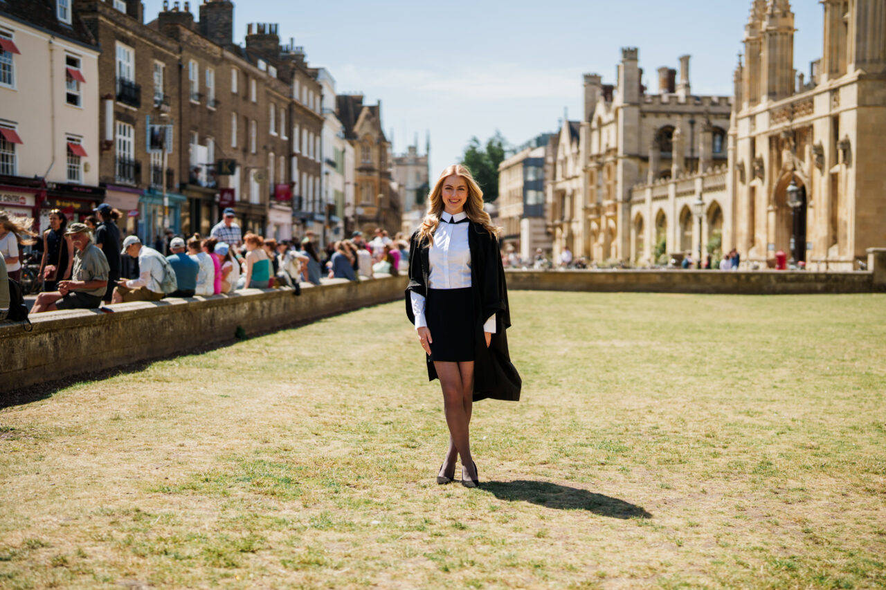 Cambridge graduation portrait of a smiling graduate in academic gown outside King's College on a sunny summer day.