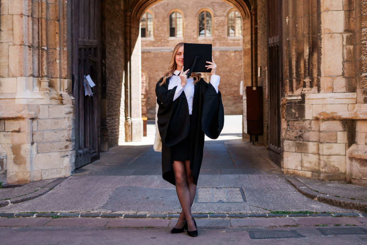 Cambridge graduation photo of a graduate playfully covering half her face with her graduation cap beneath a college archway.