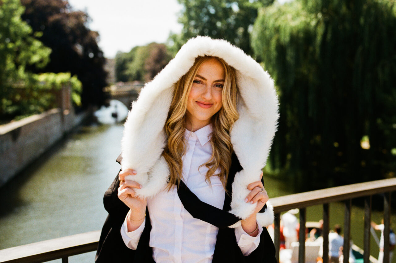 Natural Cambridge graduation portrait of a graduate by the River Cam wearing academic robes with a fur-trimmed hood.