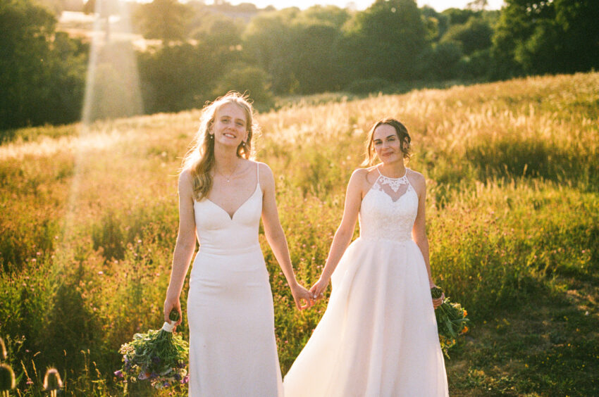 Two brides walking hand in hand through the Cambridgeshire countryside at sunset after their wedding