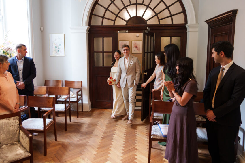 Bride and groom enter the ceremony room at Chelsea Old Town Hall hand in hand as family and friends stand to welcome them with warm smiles.