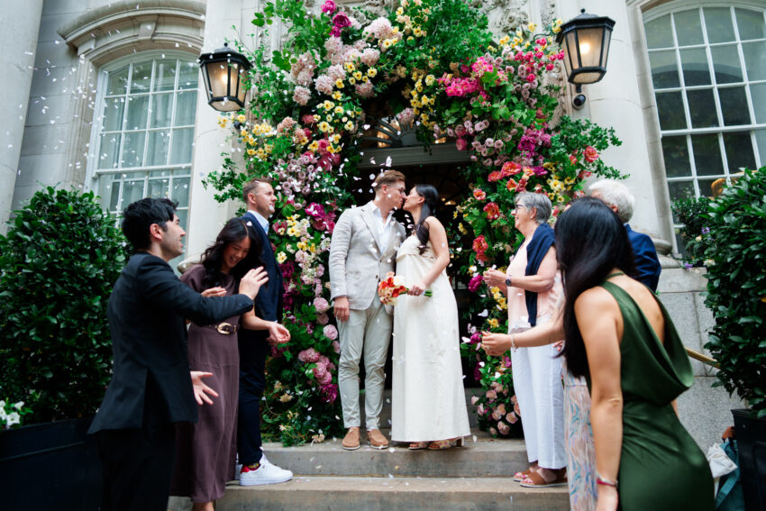Newlyweds share a kiss beneath the famous floral entrance at Chelsea Old Town Hall as guests throw confetti in celebration.