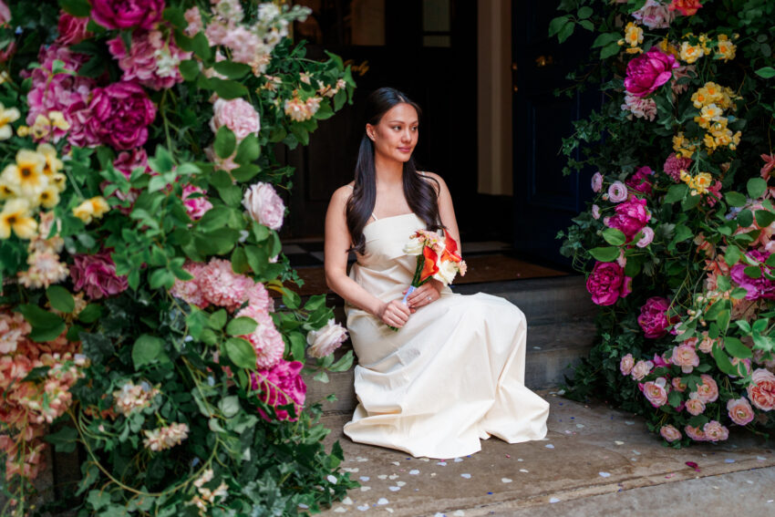 Bride seated beneath the vibrant floral installation outside Chelsea Old Town Hall, holding her bouquet during a calm, elegant portrait after the ceremony.