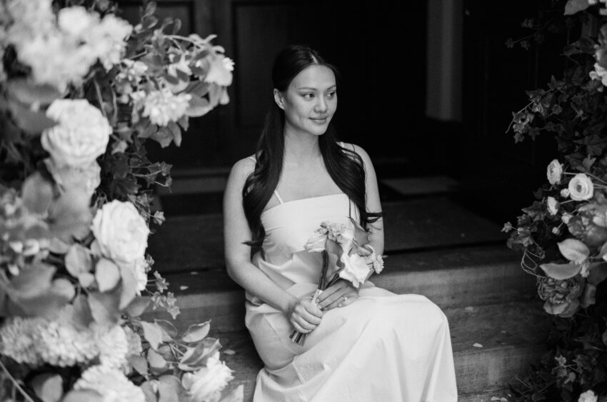 Black and white bridal portrait outside Chelsea Old Town Hall, capturing a quiet moment as the bride sits framed by abundant summer flowers.