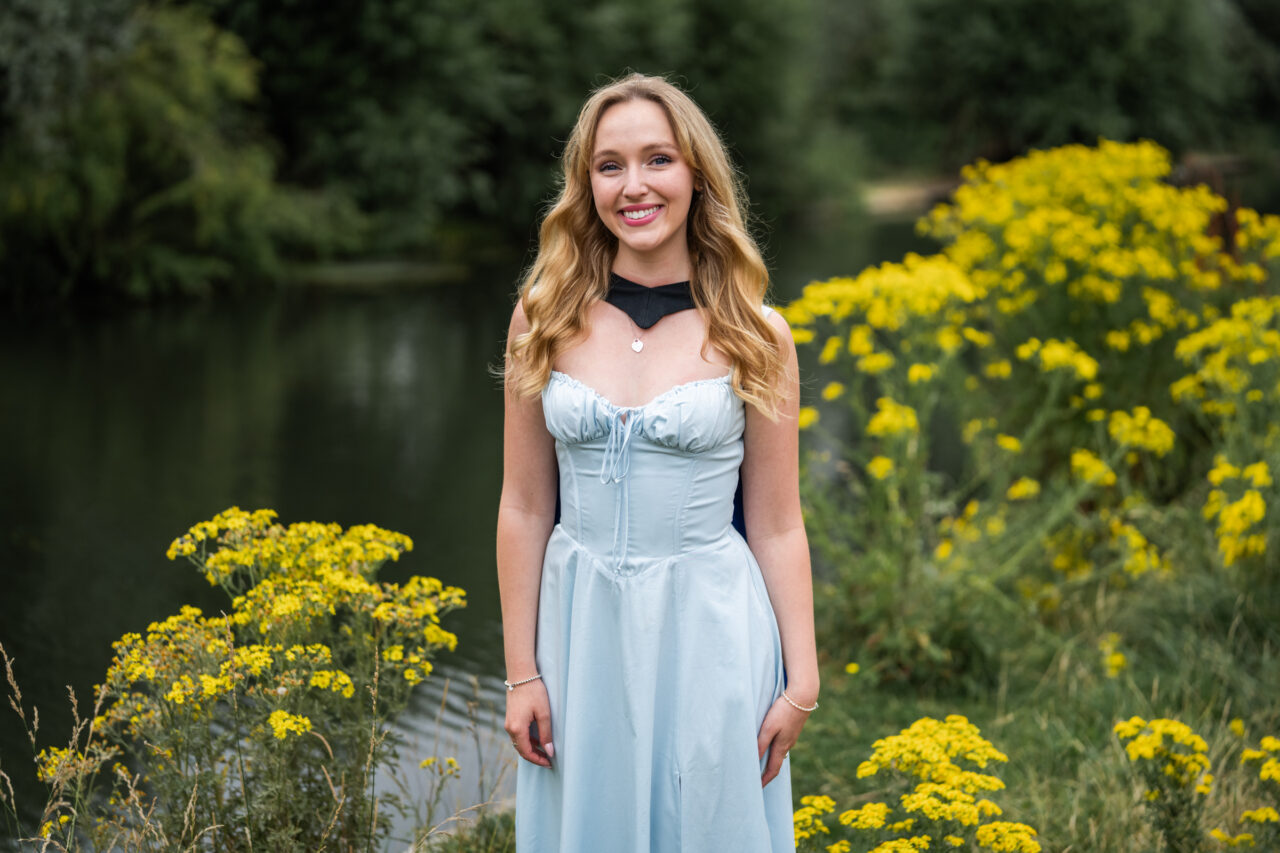 Cambridge graduate wearing graduation gown and light blue dress beside the River Cam surrounded by summer wildflowers.