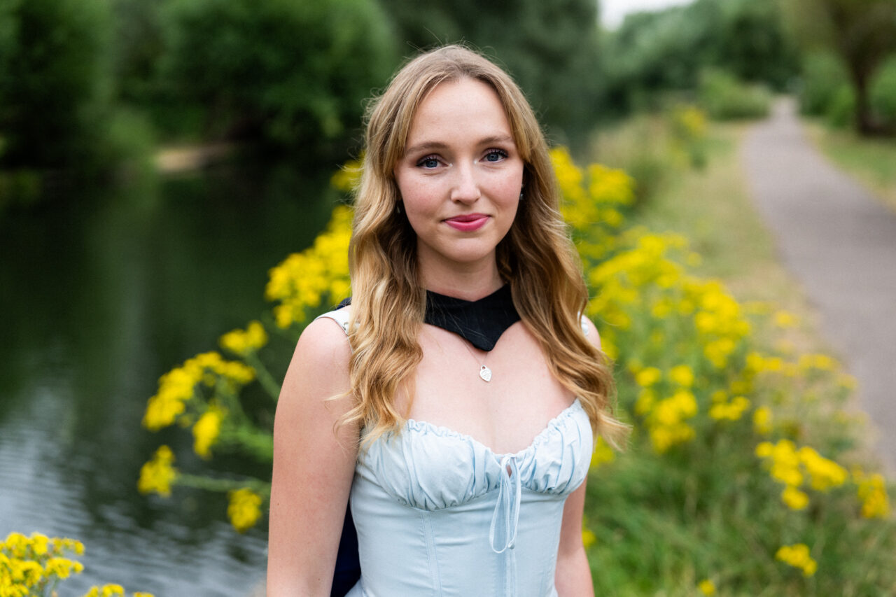 Natural graduation portrait beside the River Cam with graduation gown and yellow wildflowers at The Backs, Cambridge
