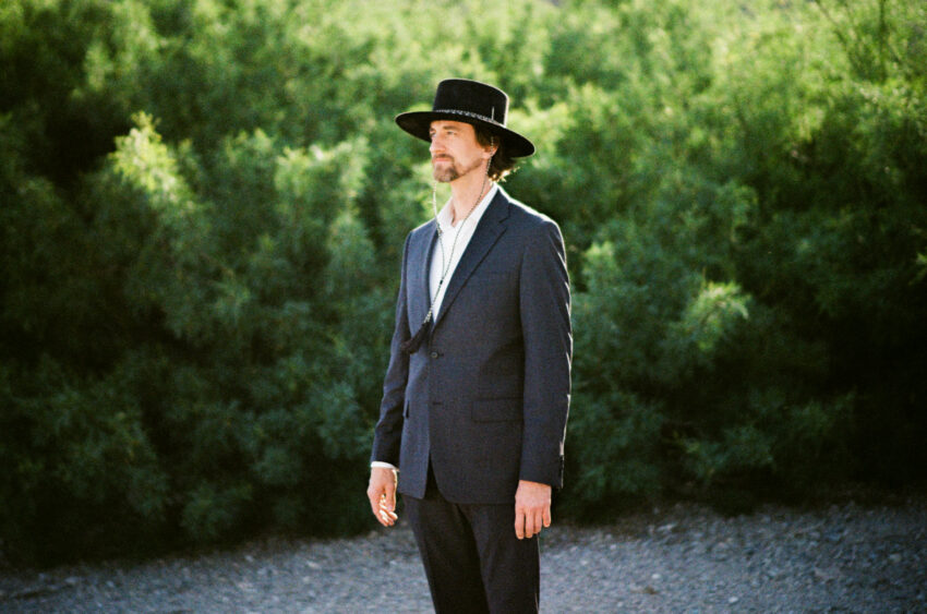 Analogue film portrait of groom wearing a black wide-brimmed hat and dark suit against lush green foliage.