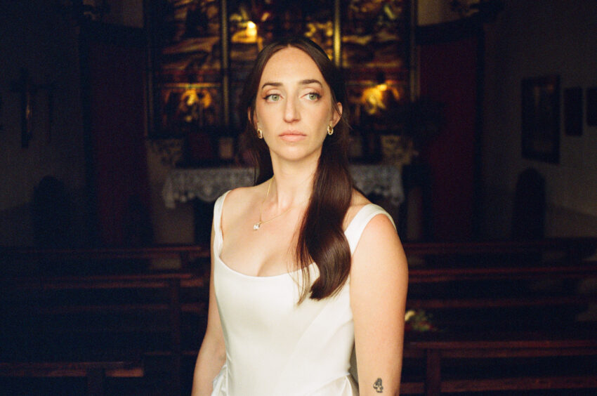 Contax T2 film portrait of a bride in a white wedding dress inside a dark Barcelona chapel, with a small arm tattoo visible.