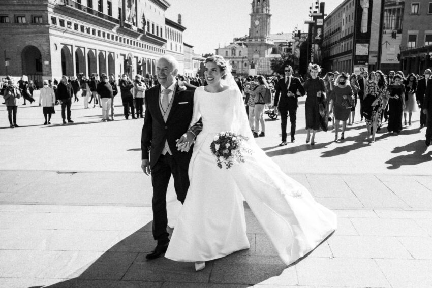 Black and white film photograph of a bride walking with her father through a sunlit Spanish square, followed by wedding guests.