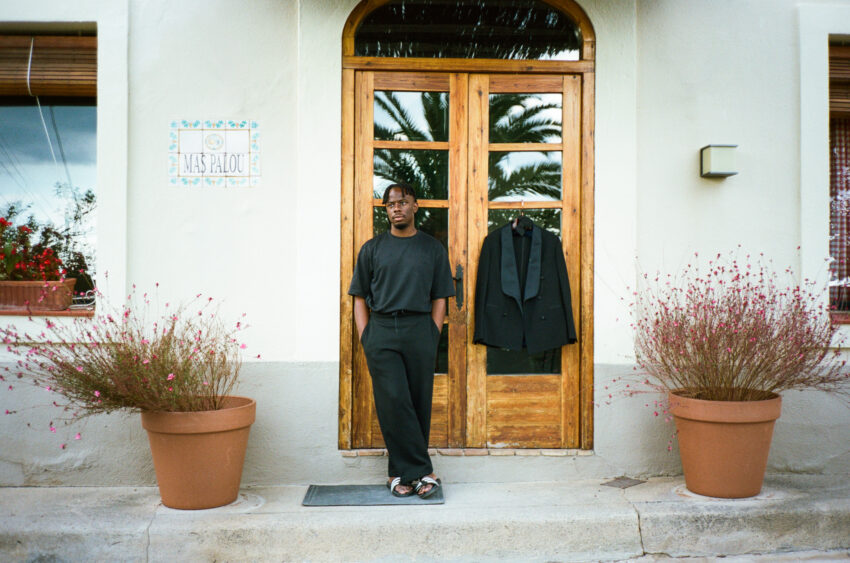 A groom in casual black clothing beside his hanging tuxedo at Mas Palou near Barcelona, photographed on 35mm film.